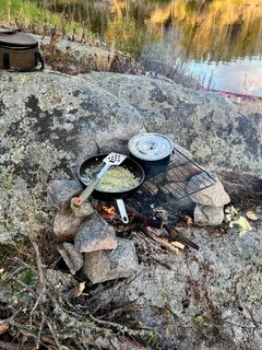A frying pan and pot on a grate over a stone firepit on granite, with the river behind