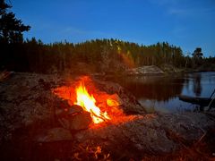 A fire burning in a rock firepit on the shoreline at dusk, pine forest across the calm river